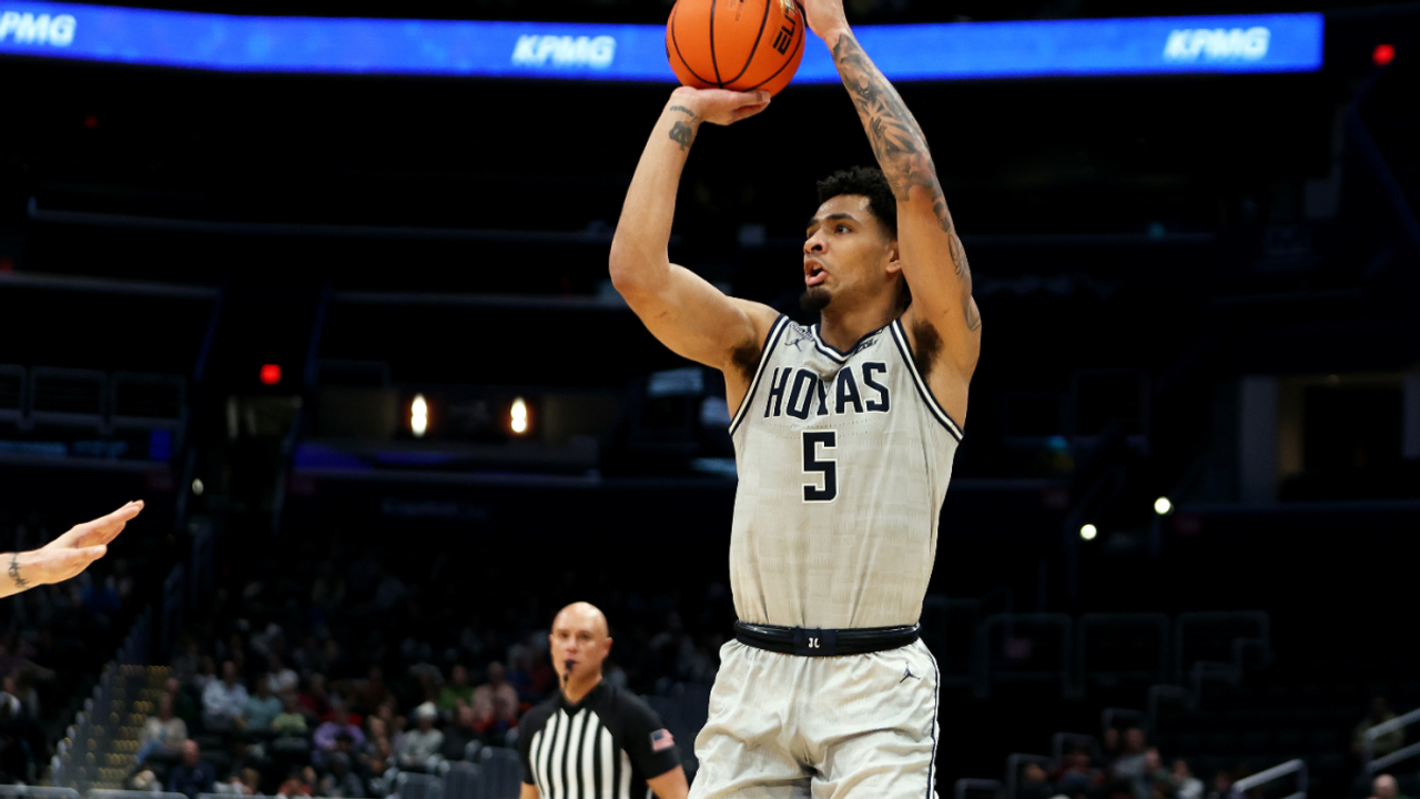 An action-packed basketball scene depicting the Georgetown Hoyas facing off against the UAlbany Great Danes. The hardwood court is illuminated, showcasing players in vibrant blue and gray jerseys, with a crowd roared in excitement. Micah Peavy, in an intense moment, is seen driving towards the basket, preparing for a layup against a defender while teammates in the background are poised for a rebound. The scoreboard above displays a dominating lead for the Hoyas, capturing the thrill of this high-scoring game. The lighting is bright, highlighting the players' expressions of determination and skill.